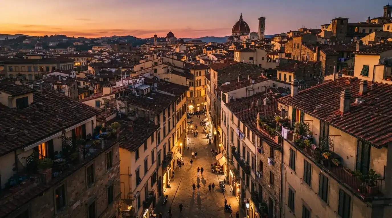 Vista dall'alto di una città italiana al tramonto, con luci che si accendono nelle finestre degli edifici lungo una via trafficata.